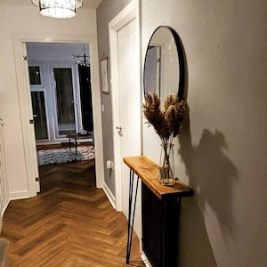 May include: A hallway with a wooden floor and grey walls. A black radiator with a wooden top and a vase of dried flowers sits against the wall. A round black mirror hangs above the radiator. A light fixture hangs from the ceiling.