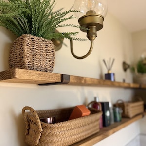 May include: Two wooden shelves with metal brackets, a wicker basket, a potted plant, and various bottles and boxes on the shelves. The shelves are mounted on a white wall with a gold light fixture above them.