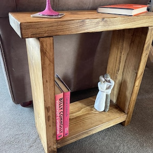 May include: A wooden side table with a single shelf. The table has a natural wood finish and is decorated with a book, a small figurine, and a glass of pink liquid.