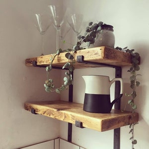 May include: Two wooden shelves with black metal brackets, a white jug with a black bottom, and a glass vase with greenery on the top shelf.