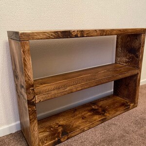 May include: A rustic wooden shelf unit with two shelves. The shelf is made of dark brown wood with a natural wood grain finish.