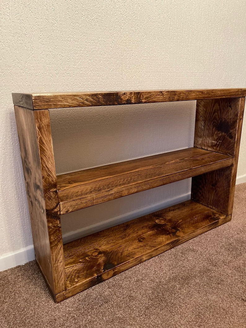 May include: A rustic wooden shelf unit with two shelves. The shelf is made of dark brown wood with a natural wood grain finish.