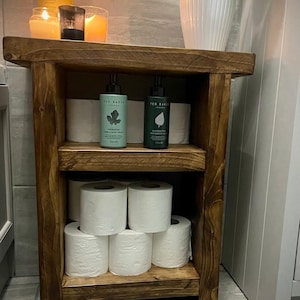 May include: A dark wooden bathroom storage unit with three shelves. The top shelf displays candles and a plant. The middle shelf holds Ted Baker products and toilet rolls. The bottom shelf has more toilet rolls and a set of scales.