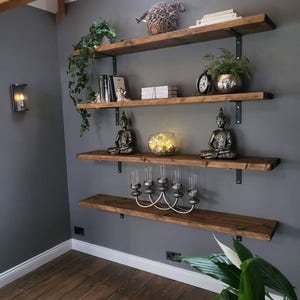 May include: A set of four wooden shelves with black metal brackets mounted on a grey wall. The shelves are decorated with plants, books, candles, and decorative items.