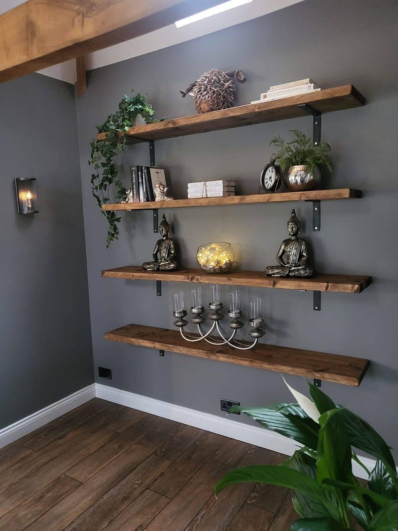 May include: A set of four wooden shelves with black metal brackets mounted on a grey wall. The shelves are decorated with plants, books, candles, and decorative items.