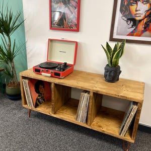 May include: A wooden record player stand with three shelves, each filled with vinyl records. The stand has hairpin legs and is made of reclaimed wood. A red record player sits on top of the stand.
