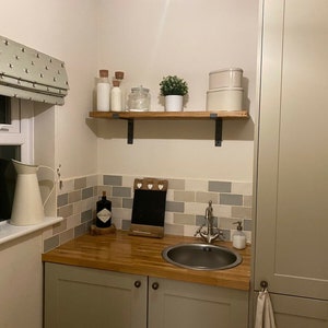 May include: A small kitchen or utility room with a wooden countertop, a stainless steel sink, and a white cabinet with a towel hanging from the handle. There is a shelf above the countertop with jars and a small plant. The walls are tiled in a light gray and white pattern.