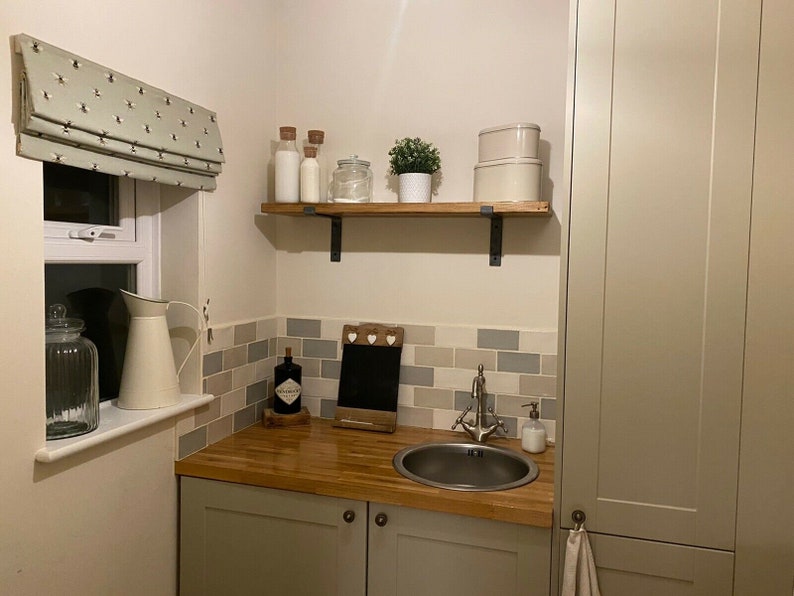 May include: A small kitchen or utility room with a wooden countertop, a stainless steel sink, and a white cabinet with a towel hanging from the handle. There is a shelf above the countertop with jars and a small plant. The walls are tiled in a light gray and white pattern.