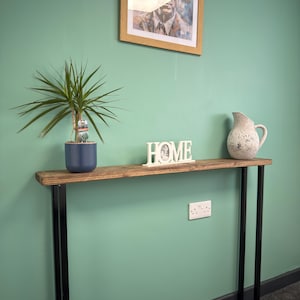 May include: A wooden console table with black metal legs, holding a framed portrait, a potted plant, a "HOME" sign, and a speckled ceramic jug. The table is against a teal wall.