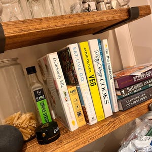 May include: A wooden shelf with a variety of cookbooks, including "Deliciously Ella", "Eat. Live. Go.", "Jamie Oliver Veg", "Mary Berry Cooks", and "Itsu the Cookbook". There is also a bottle of simple syrup, a jar of pasta, and a black round object on the shelf.