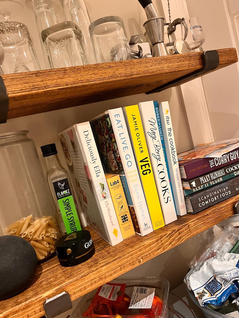 May include: A wooden shelf with a variety of cookbooks, including "Deliciously Ella", "Eat. Live. Go.", "Jamie Oliver Veg", "Mary Berry Cooks", and "Itsu the Cookbook". There is also a bottle of simple syrup, a jar of pasta, and a black round object on the shelf.