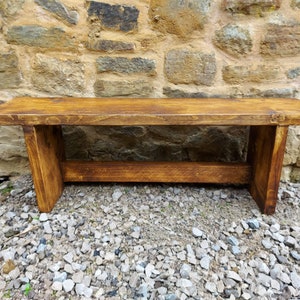 May include: A rustic wooden bench with a natural brown stain finish. The bench has a simple design with a single plank for the seat and two sturdy legs.