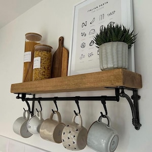 May include: A rustic wooden shelf with black metal brackets and a black metal rail with hooks. The shelf is holding two glass jars filled with pasta, a framed print with the text "Italian Pasta The Guide", and a potted succulent plant. There are six mugs hanging from the hooks.