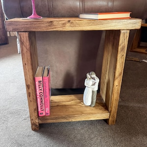 A wooden side table with a pink wine glass and a book on it, along with a small figurine of a couple.