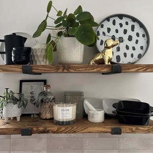 a wooden shelf with various decorative items and plants, including a black and white polka dot plate, a gold dog figurine, and a white vase with green leaves. The shelf also holds a black teapot, a white candle, and a white bowl.