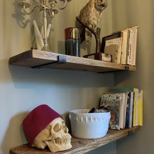 Puede incluir: Dos estantes de madera con soportes de metal negro, un estante est&aacute; vac&iacute;o y el otro tiene un taz&oacute;n blanco, un sombrero rojo y una calavera blanca. El estante superior tiene un b&uacute;ho de peluche, un jarr&oacute;n rojo y azul y una pila de libros.