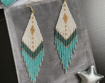 May include: A pair of long, beaded earrings with a geometric design in white, gold, and teal blue. The earrings are hanging from gold hooks.