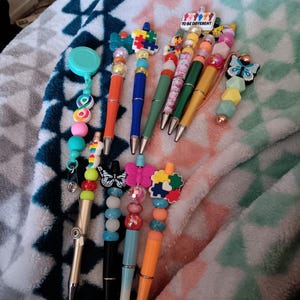 May include: A collection of decorative pens with colorful beads and charms. Some pens feature butterfly, puzzle piece, and "TO BE DIFFERENT" designs. The pens have orange, blue, green, and black barrels. The pens are arranged on a patterned blanket.