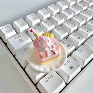 May include: A miniature dessert, resembling a cake with ice cream, sits on a small white plate atop a white keyboard. The cake is pink, yellow, and white, with a bow and a small ice cream cone. The keyboard keys are white.