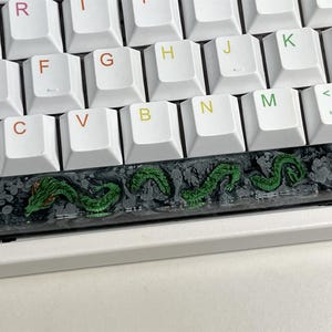 May include: A black and green keyboard keycap with a dragon design. The keycap is made of resin and has a glossy finish.