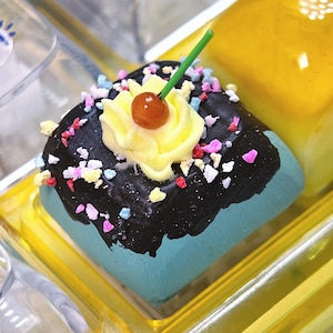 May include: A miniature dessert with a blue base, topped with dark chocolate frosting, yellow frosting, a cherry, and colorful sprinkles. The dessert is in a clear plastic container with a yellow base. The image is a close-up shot.