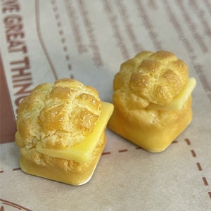 May include: Two miniature, yellow, bread-like pastries with a yellow, butter-like filling. The pastries are on a brown paper surface.