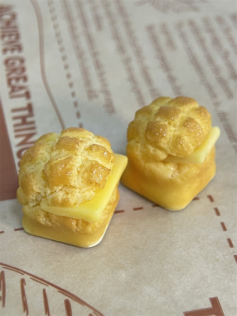 May include: Two miniature, yellow, bread-like pastries with a yellow, butter-like filling. The pastries are on a brown paper surface.