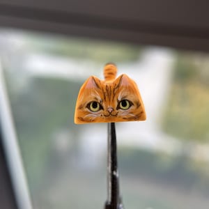 May include: A detailed, orange cat face keycap with yellow eyes and a striped tail. The keycap is held by a black tool, with a blurred background of a window and greenery.