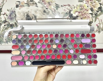 May include: A white and silver mechanical keyboard with round keys. The keys are translucent and have different coloured lipstick shades underneath. The keyboard has a row of keys labelled "Esc", "F1", "Tab", "CapsLk", "Shift", "Ctrl", "Alt", "Fn", and "Alt".
