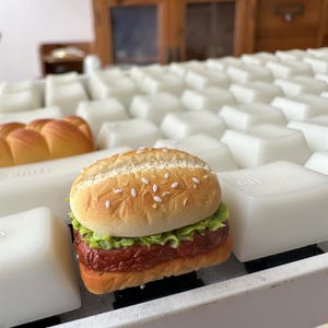 Peut inclure: Un hamburger miniature avec un pain au sésame, de la laitue et un steak haché, posé sur un clavier blanc.