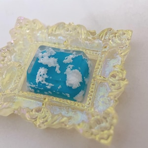 Könnte beinhalten: Ein kleines, blaues Harzstück mit weißen Wirbeln, das an Wolken erinnert, wird in einem gelben, verzierten Rahmen präsentiert.