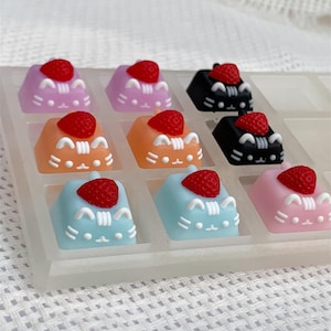 Può includere: Nove tasti colorati con un designo a forma di gatto e una fragola rossa in cima. I tasti sono disposti in una griglia 3x3 su una superficie bianca.