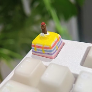 May include: A square mechanical keyboard keycap shaped like a multi-layered cake with horizontal stripes in pink, blue, and white. The top is covered in yellow icing, a white dollop of frosting, and a small brown cylindrical chocolate garnish.