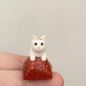 May include: A small white ceramic cat figurine sitting on a red ceramic strawberry. The cat has black eyes and a white face.
