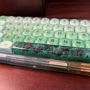 Könnte beinhalten: Mechanische Tastatur mit grün-weißen Keycaps. Die Leertaste ist aus transparentem Harz mit dunklem Design. Die Tastatur liegt auf einer Holzoberfläche.