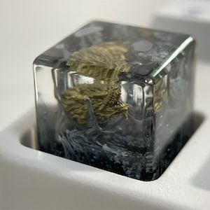May include: A clear resin keycap with a gold-colored design inside. The keycap is in a white keycap holder.