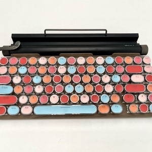 May include: A retro-style keyboard with round keys in various shades of pink, red, and blue. The keys have white lettering and include function keys, number keys, and letter keys. The keyboard has a wooden frame and a black metal bar on top.
