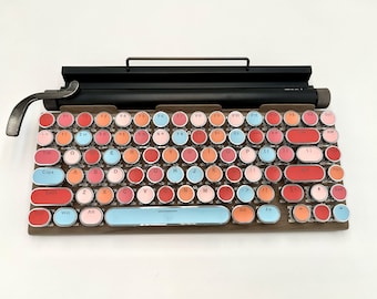 Op de afbeelding: Een retro-stijl toetsenbord met ronde toetsen in verschillende tinten roze, rood en blauw. De toetsen hebben witte letters en omvatten functietoetsen, cijfertoetsen en lettertoetsen. Het toetsenbord heeft een houten frame en een zwarte metalen balk bovenop.
