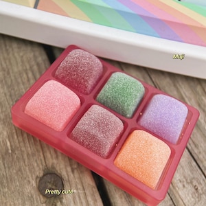 以下が含まれることがあります： 赤いプラスチック製のトレイに、6つの正方形の区画があり、カラフルな砂糖菓子で満たされています。キャンディーはグリッドパターンで配置されており、ざらざらした質感です。トレイは木製の表面の上に置かれています。