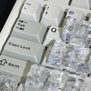 Puede incluir: Primer plano de un teclado de ordenador con teclas blanquecinas. Las teclas incluyen símbolos y letras como "Tab", "Shift", "Caps Lock", "Q", "1" y "!". Formas claras y texturizadas, parecidas al hielo, se colocan encima de algunas teclas.