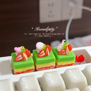 Op de afbeelding: Drie vierkante mechanische toetsenbordtoetsen in de vorm van miniatuur laagjescakes. Elke cake heeft een groene bovenlaag, een gele tussenlaag en een roze onderlaag, afgewerkt met kleine aardbeiplakjes en roze dierfiguren.