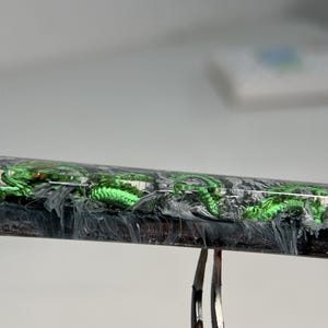 May include: A close-up of a black and white resin pen with green glitter embedded in the resin. The pen is held in place by a pair of tweezers.