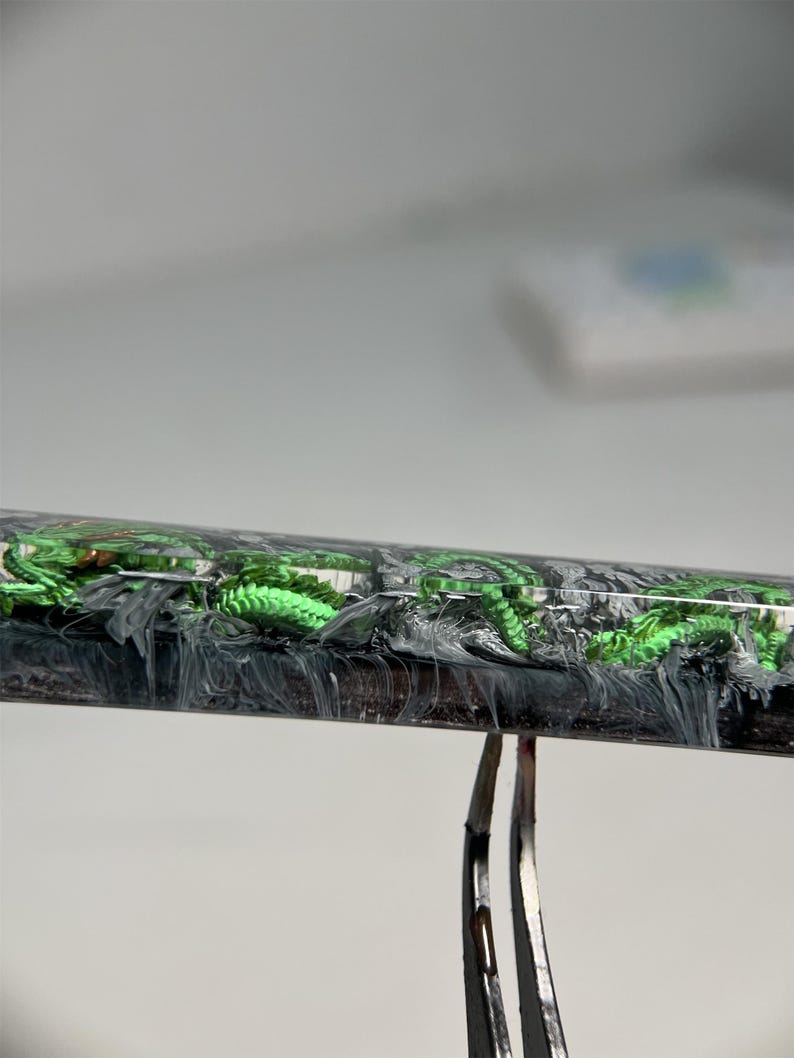 May include: A close-up of a black and white resin pen with green glitter embedded in the resin. The pen is held in place by a pair of tweezers.