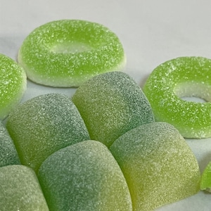 Op de afbeelding: Assortiment van groene gummy snoepjes in verschillende vormen, waaronder ringen en vierkanten. De snoepjes hebben een korrelige textuur en een lichtgroene kleur.