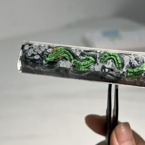 May include: A clear resin cylinder with a green and gold dragon design. The dragon is curled up inside the cylinder, which is held by tweezers.