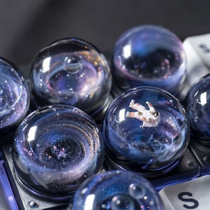 May include: A set of six clear glass keycaps with a galaxy theme. Each keycap features a different design, including a swirling galaxy, a nebula, and an astronaut floating in space.