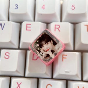 Puede incluir: Un tapón de tecla de resina rosa y transparente con una foto de una mujer con el pelo largo y negro. El tapón de tecla está en un teclado blanco con letras rosas.