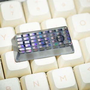 May include: A custom keyboard keycap with a clear, iridescent design, resting on a cream-colored keyboard. The keycap has the word "LED" visible. The keycap is a unique accessory for mechanical keyboards.