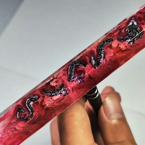 May include: A clear rectangular block of resin with a swirling red and pink pattern. Embedded in the resin are multiple black and purple dragon-like figures.