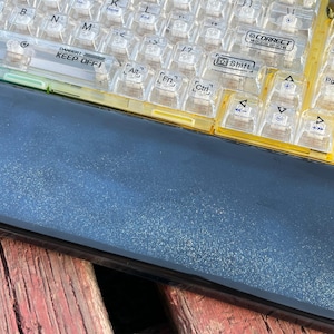 K&ouml;nnte beinhalten: Eine gelbe und transparente Tastatur aus Kunststoff mit schwarzen Beschriftungen. Die Tastatur hat ein schwarzes und silbernes Geh&auml;use mit einem Warnhinweis, der "DANGER! KEEP OFF!" lautet. Die Tastatur liegt auf einer Holzoberfl&auml;che.